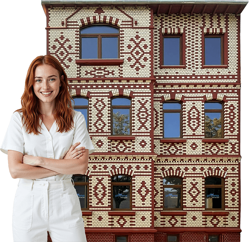 Eine lächelnde Frau mit langen roten Haaren und weißer Kleidung steht vor einem Phoenix Homecare-Gebäude mit verziertem Fassadenmuster.